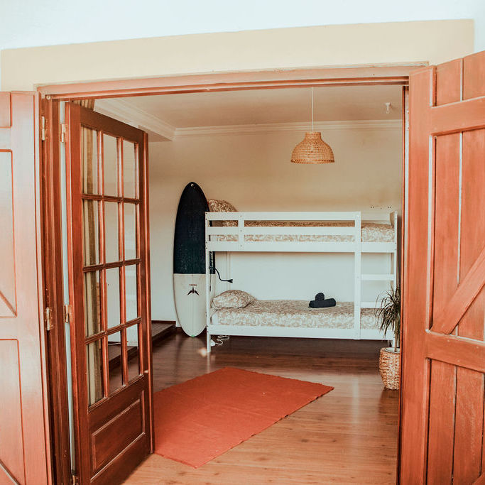 Shared room with white bunk beds, ample space, and natural light pouring in. A surfboard leans against the wall, adding a touch of adventure to the inviting and spacious accommodation.