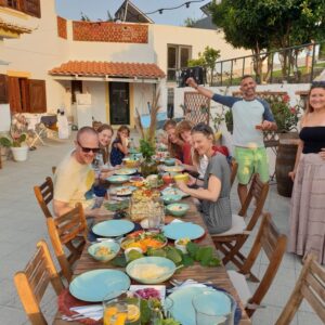 A joyful group of people, smiles on their faces, ready to gather for dinner outdoors. The table is adorned with plates, decorations, and an array of delicious dishes. The warm glow of the sunset lights up the scene, creating a festive and inviting atmosphere.