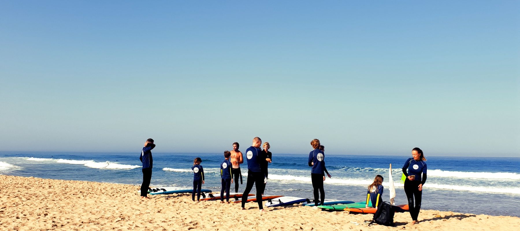 A joyful family group participating in surf lessons at Praia da Areia Branca, Far End Surf House. The beach setting, with waves in the background, captures the excitement and camaraderie of a memorable surfing experience for all ages.
