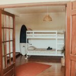 Looking into a shared room from outside, featuring a white bunk bed and a blue-and-white glass-fiber surfboard leaning against the wall. The doors are adorned with small square glass panels, allowing glimpses into the inviting space