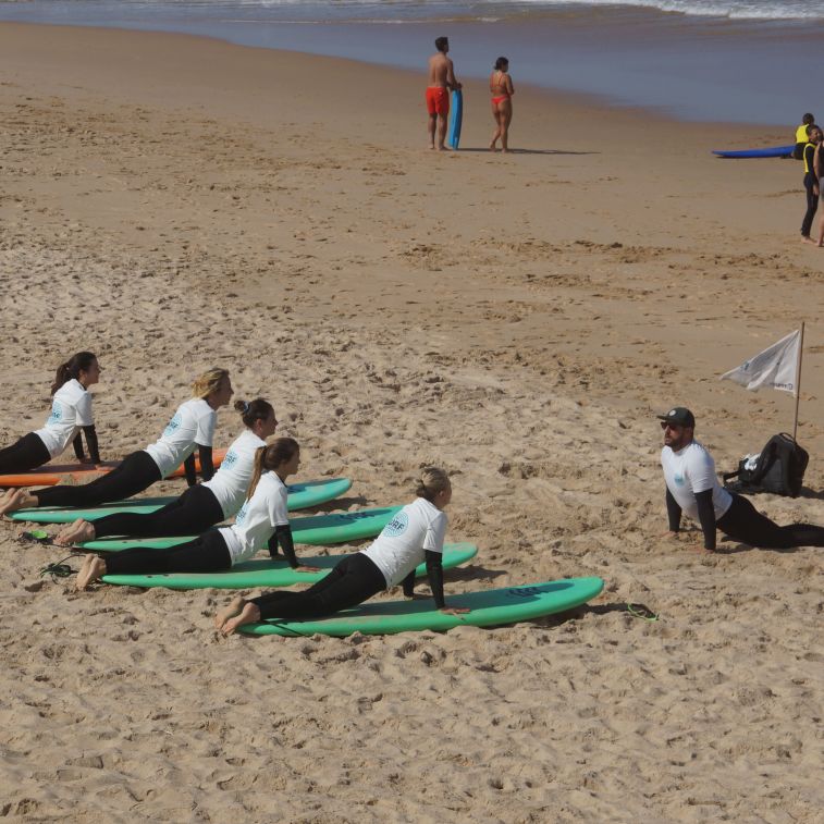 People on the beach wearing black wetsuits and white lycra, laying on surfboards, receiving instructions from surf teacher Pedro.