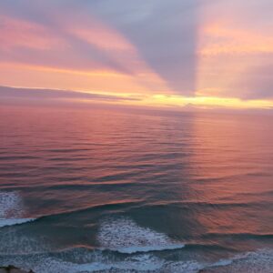 Vibrant hues paint the sky as the sun sets over the vast ocean, viewed from the majestic vantage point atop a cliff. Waves dance below, reflecting the warm and golden glow, creating a serene and breathtaking scene