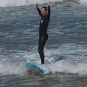 A girl in a wetsuit celebrating catching waves, arms raised in triumph with a joyful smile on her face as she learns to surf.