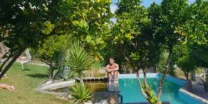A boy sits at the edge of a saltwater pool with a simple pond at the far end, surrounded by trees and nature at The Far End Surf House.