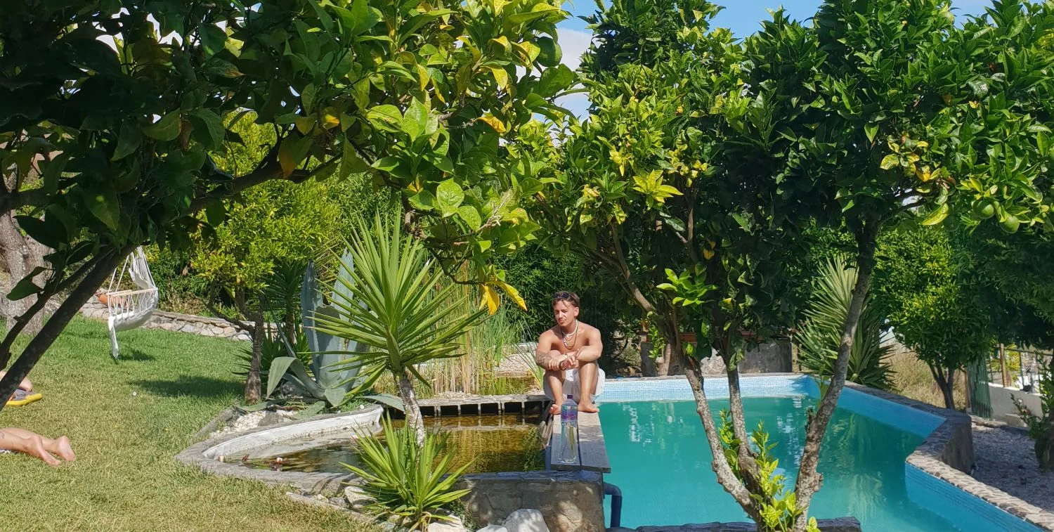 A boy sits at the edge of a saltwater pool with a simple pond at the far end, surrounded by trees and nature at The Far End Surf House.