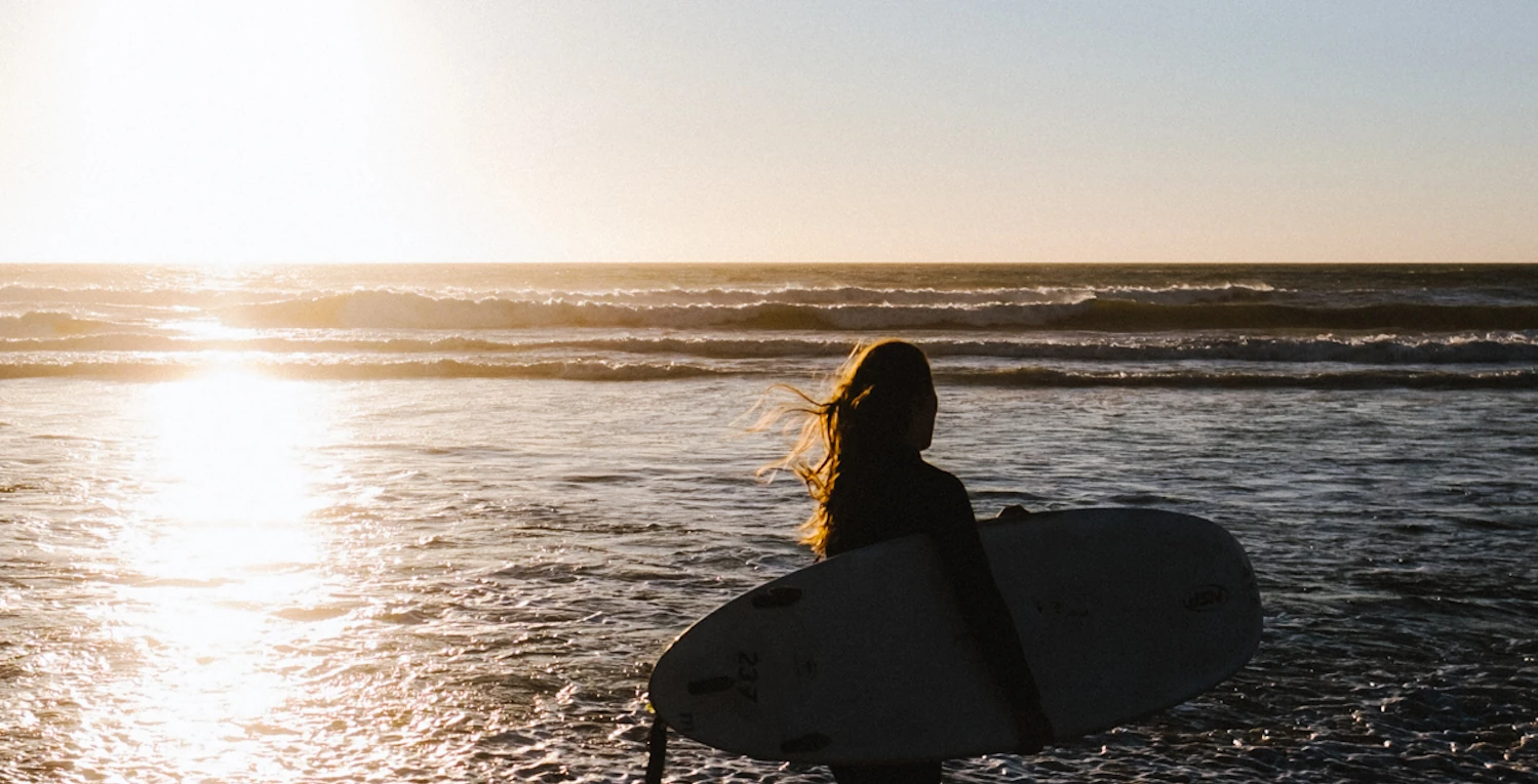A girl holding a surfboard walks toward the ocean at sunset in Portugal, seen from behind.