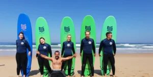 Five students stand on the beach with surfboards behind their backs, while the surf instructor kneels in front of them in Portugal.