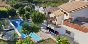 View of a villa with a swimming pool, surrounded by a green garden and countryside in the background.