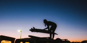 Silhouette of a girl loading surfboards on top of a car at sunset, with purple-colored sky.